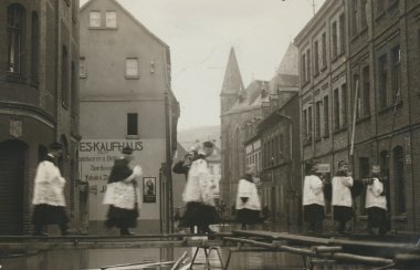 Eine Straße in einer Stadt mit Kirche im Hintergrund. Mehrere Personen in heller Kleidung gehen über provisorische Stege durch eine überschwemmte Straße; Gebäude mit Geschäften säumen den Weg.