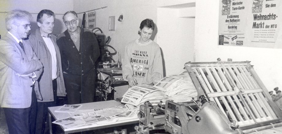 Schwarz-Weiß-Foto: Mehrere Erwachsene und ein Jugendlicher stehen in einem Raum neben einer großen technischen Maschine mit Rollen und Hebeln; sie betrachten die Anlage, im Hintergrund hängen Plakate an der Wand.