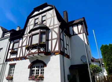 Traditionelles Fachwerkhaus mit weißer Fassade und dunklen Holzbalken, Blumen an den Fenstern und blauem Himmel im Hintergrund.