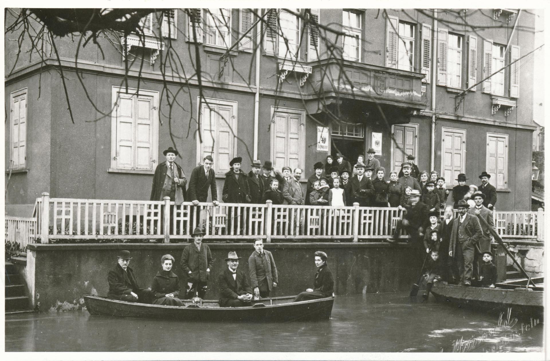 Historische Schwarz-Weiß-Aufnahme eines überfluteten Gebäudes. Zahlreiche Menschen stehen auf einer erhöhten Veranda und beobachten die Hochwassersituation, während mehrere Personen in einem Ruderboot vor dem Haus auf dem überfluteten Vorplatz sitzen. Im Vordergrund ist das Wasser zu sehen, rechts ein weiteres Boot mit Menschen.