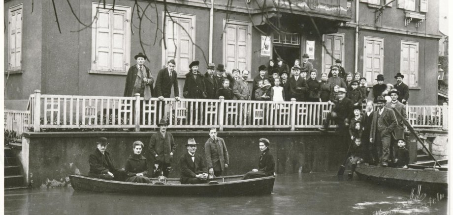 Historische Schwarz-Weiß-Aufnahme eines überfluteten Gebäudes. Zahlreiche Menschen stehen auf einer erhöhten Veranda und beobachten die Hochwassersituation, während mehrere Personen in einem Ruderboot vor dem Haus auf dem überfluteten Vorplatz sitzen. Im Vordergrund ist das Wasser zu sehen, rechts ein weiteres Boot mit Menschen.