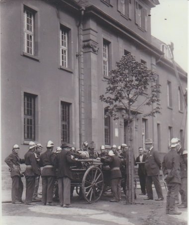Historische Schwarz-Weiß-Aufnahme eines Gebäudes, vor dem eine Gruppe von Feuerwehrleuten mit einem handgezogenen Löschwagen im Einsatz steht. Einige Einsatzkräfte bedienen das Gerät, während weitere Personen das Geschehen beobachten. Auf dem Dach ist ein Feuerwehrmann auf einer Leiter zu erkennen.