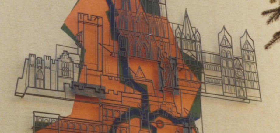 Großes Wandkunstwerk an einer hellen Gebäudefassade. Die Gestaltung zeigt stilisierte Umrisse mehrerer historischer Bauwerke und eines Denkmals, kombiniert mit orangefarbenen und grünen Flächen. Im unteren Bereich ist der Schriftzug „Ernst Wagner“ zu erkennen. Vor der Wand ragen die Zweige eines Strauchs ins Bild.
