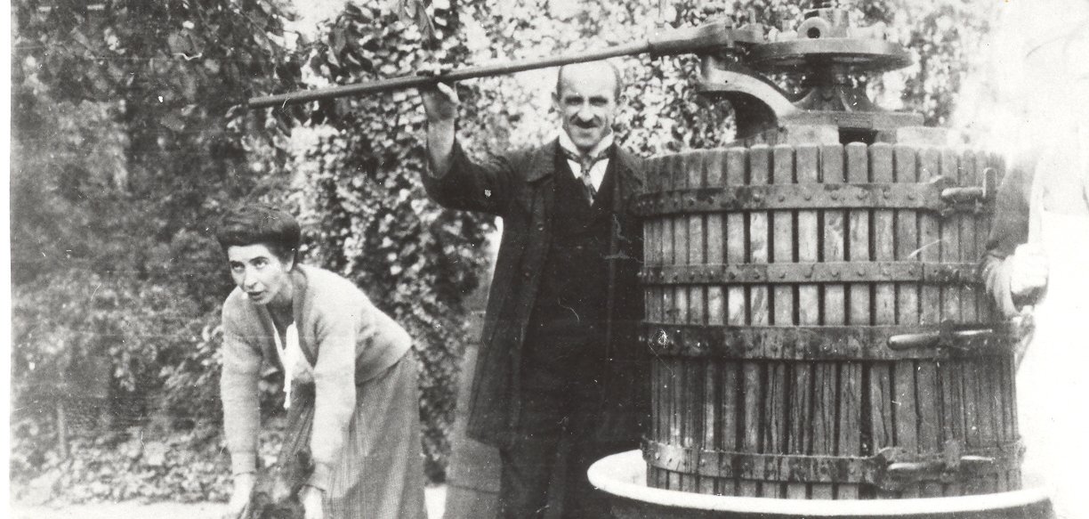 Historische Schwarz-Weiß-Aufnahme einer Weinlese: Eine Person bedient eine große hölzerne Spindelkelter mit langem Hebelarm, während eine zweite Person sich mit einem Behälter daneben vorbeugt. Die massive Weinpresse steht im Freien unter Bäumen und ist von Eimern und weiteren Arbeitsutensilien umgeben.