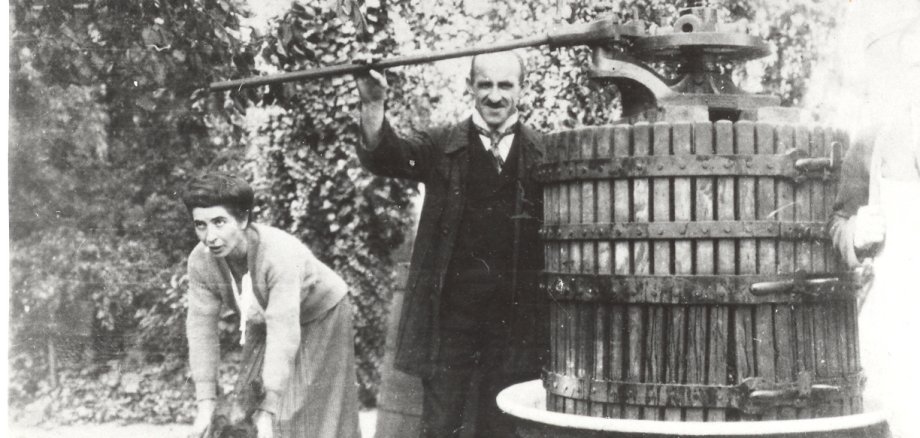 Historische Schwarz-Weiß-Aufnahme einer Weinlese: Eine Person bedient eine große hölzerne Spindelkelter mit langem Hebelarm, während eine zweite Person sich mit einem Behälter daneben vorbeugt. Die massive Weinpresse steht im Freien unter Bäumen und ist von Eimern und weiteren Arbeitsutensilien umgeben.