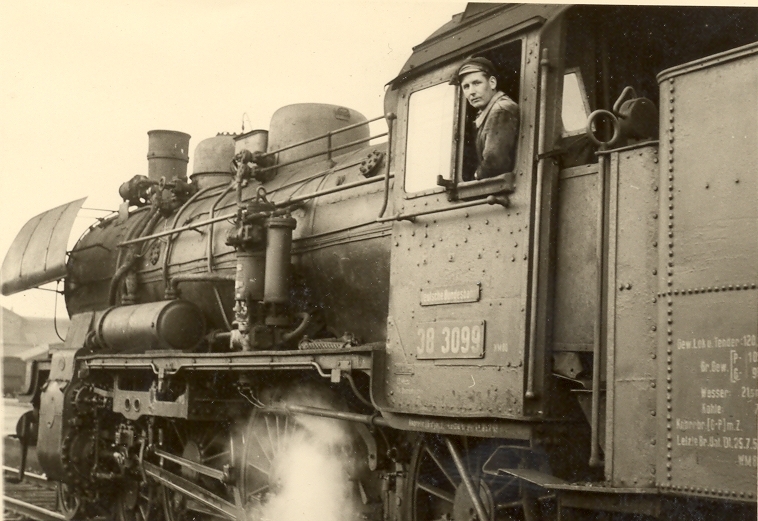 Historische Schwarz-Weiß-Aufnahme einer Dampflokomotive. Ein Lokführer schaut aus dem Fenster des Führerstands in die Kamera. Im Vordergrund sind die Räder, das Gestänge und aufsteigender Dampf zu sehen.