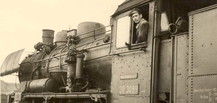 Lahnstein hat Geschichte Historische Schwarz-Weiß-Aufnahme einer Dampflokomotive. Ein Lokführer schaut aus dem Fenster des Führerstands in die Kamera. Im Vordergrund sind die Räder, das Gestänge und aufsteigender Dampf zu sehen.