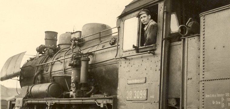 Lahnstein hat Geschichte Historische Schwarz-Weiß-Aufnahme einer Dampflokomotive. Ein Lokführer schaut aus dem Fenster des Führerstands in die Kamera. Im Vordergrund sind die Räder, das Gestänge und aufsteigender Dampf zu sehen.
