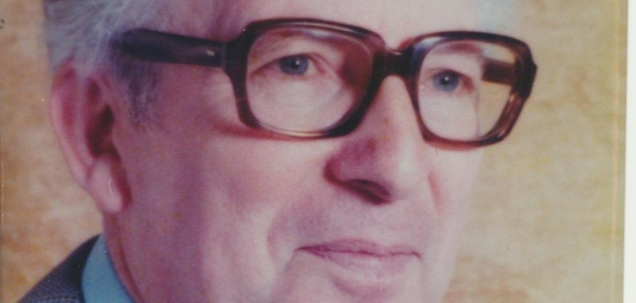 Porträtaufnahme eines älteren Mannes vor einem hellbraunen, einfarbigen Hintergrund. Er hat kurze, nach hinten gekämmte grau-weiße Haare und trägt eine große, dunkel gerahmte Brille. Bekleidet ist er mit einem hellblauen Hemd, einer dunklen Krawatte und einem grau karierten Sakko. Sein Oberkörper ist leicht seitlich ausgerichtet, während sein Blick nach vorne und etwas zur Seite gerichtet ist.