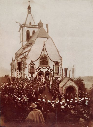 Allerheiligenbergkapelle 1901 Historische Sepiaaufnahme der festlich geschmückten Allerheiligenbergkapelle in Lahnstein. Vor dem Eingang haben sich zahlreiche Menschen zur feierlichen Einweihung versammelt. Fahnen, Girlanden und eine große Menschenmenge prägen das Bild.