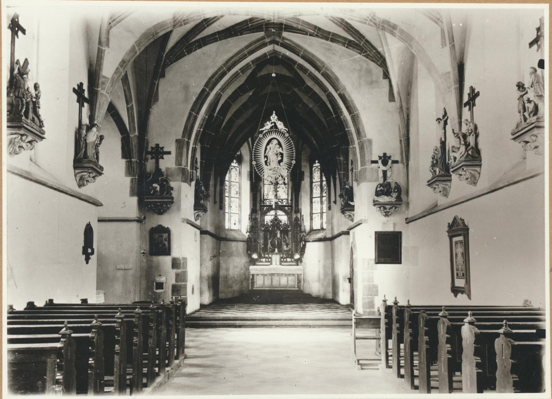 Historische Schwarz-Weiß-Aufnahme des Innenraums der Allerheiligenbergkapelle in Lahnstein. Der Blick führt zwischen den hölzernen Kirchenbänken zum reich verzierten Altar mit einer Marienfigur. Spitzbogengewölbe, hohe Kirchenfenster und Kreuzwegstationen an den Seitenwänden prägen den Kirchenraum.