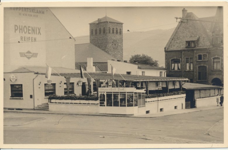 Historische Schwarz-Weiß-Aufnahme einer Straßenkreuzung in Lahnstein mit einem Gebäude, an das eine große, teilweise überdachte Außenterrasse angeschlossen ist. Die Terrasse ist von einer niedrigen weißen Mauer mit zahlreichen Blumenkästen eingefasst. An ihrer Vorderseite befindet sich ein verglaster Eingangsbereich mit der Aufschrift „Blumenhalle Koch“. Mehrere Fahnen sind an schräg aufgestellten Fahnenstangen befestigt. Links erhebt sich eine große helle Hauswand mit den Werbeaufschriften „Ruppertsklamm – 30 Min. von hier“ und „Phoenix Reifen“. Dahinter ist ein viereckiger Kirchturm mit Uhr zu erkennen. Rechts steht ein mehrgeschossiges Backsteingebäude mit hohen Fenstern und einem steilen Dach. Im Vordergrund verläuft eine breite, nahezu menschenleere Straße. Das Foto besitzt einen hellen, leicht vergilbten Rand.
