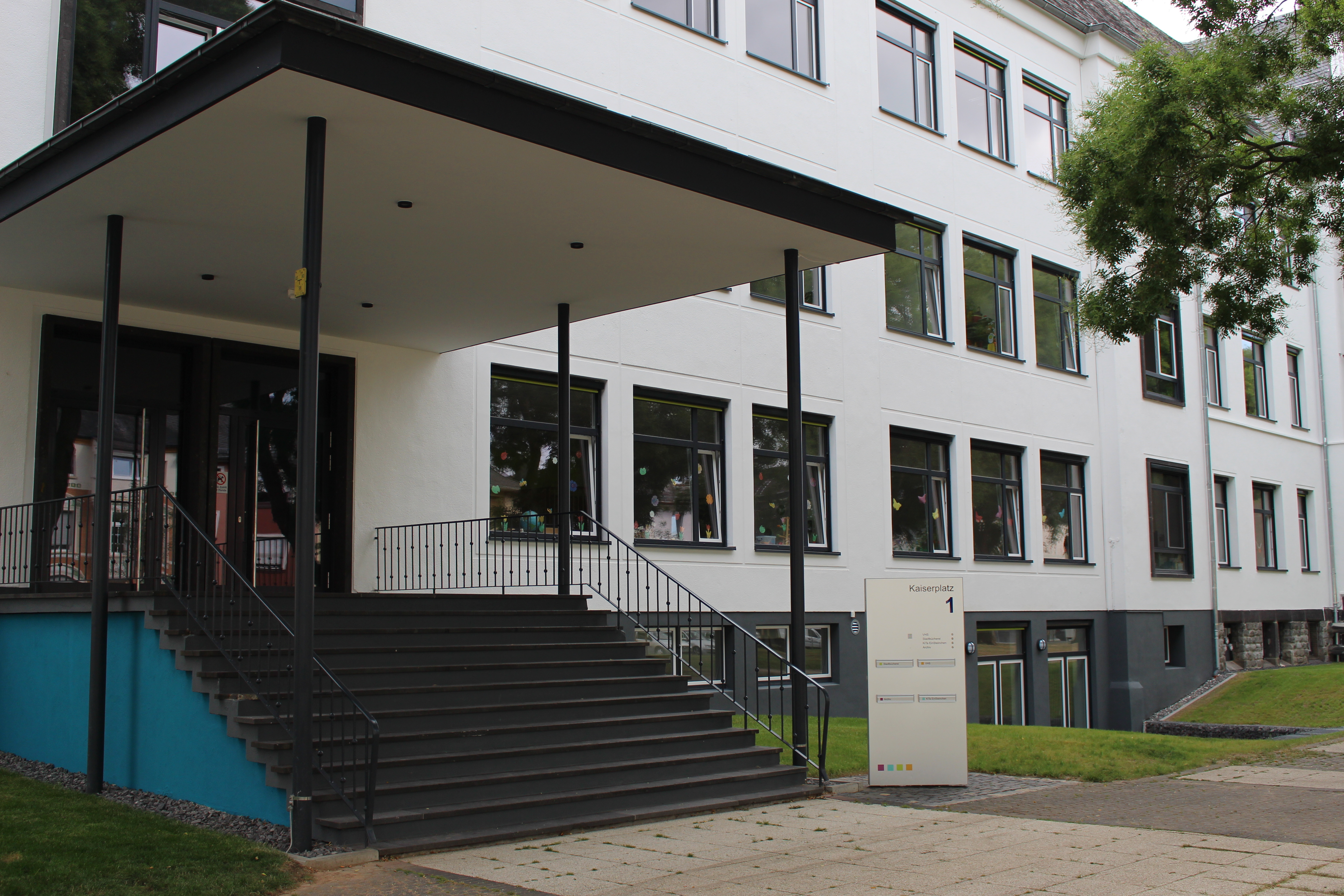 Ein hell verputztes, mehrstöckiges Gebäude mit zahlreichen großen Fenstern. Im Vordergrund führt eine breite Treppe mit Geländern zu einem überdachten Haupteingang. Vor dem Gebäude befinden sich Rasenflächen und Bäume. Rechts neben der Treppe steht eine Stele mit der Aufschrift „Kaiserplatz 1“.