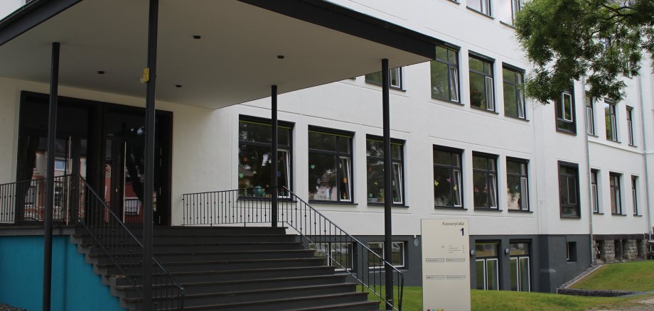 Stadtarchiv Lahnstein Ein hell verputztes, mehrstöckiges Gebäude mit zahlreichen großen Fenstern. Im Vordergrund führt eine breite Treppe mit Geländern zu einem überdachten Haupteingang. Vor dem Gebäude befinden sich Rasenflächen und Bäume. Rechts neben der Treppe steht eine Stele mit der Aufschrift „Kaiserplatz 1“.