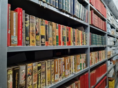 Stadtarchiv Lahnstein Mehrere dicht gefüllte Metallregale mit Büchern in einem Magazinraum. In den Regalen stehen unterschiedlich große Bücher mit farbigen Einbänden, darunter viele ältere Ausgaben und gebundene Zeitschriften. Rechts sind zusätzlich Archivkartons und weitere Bände untergebracht. Der Blick verläuft entlang der Regalreihen.