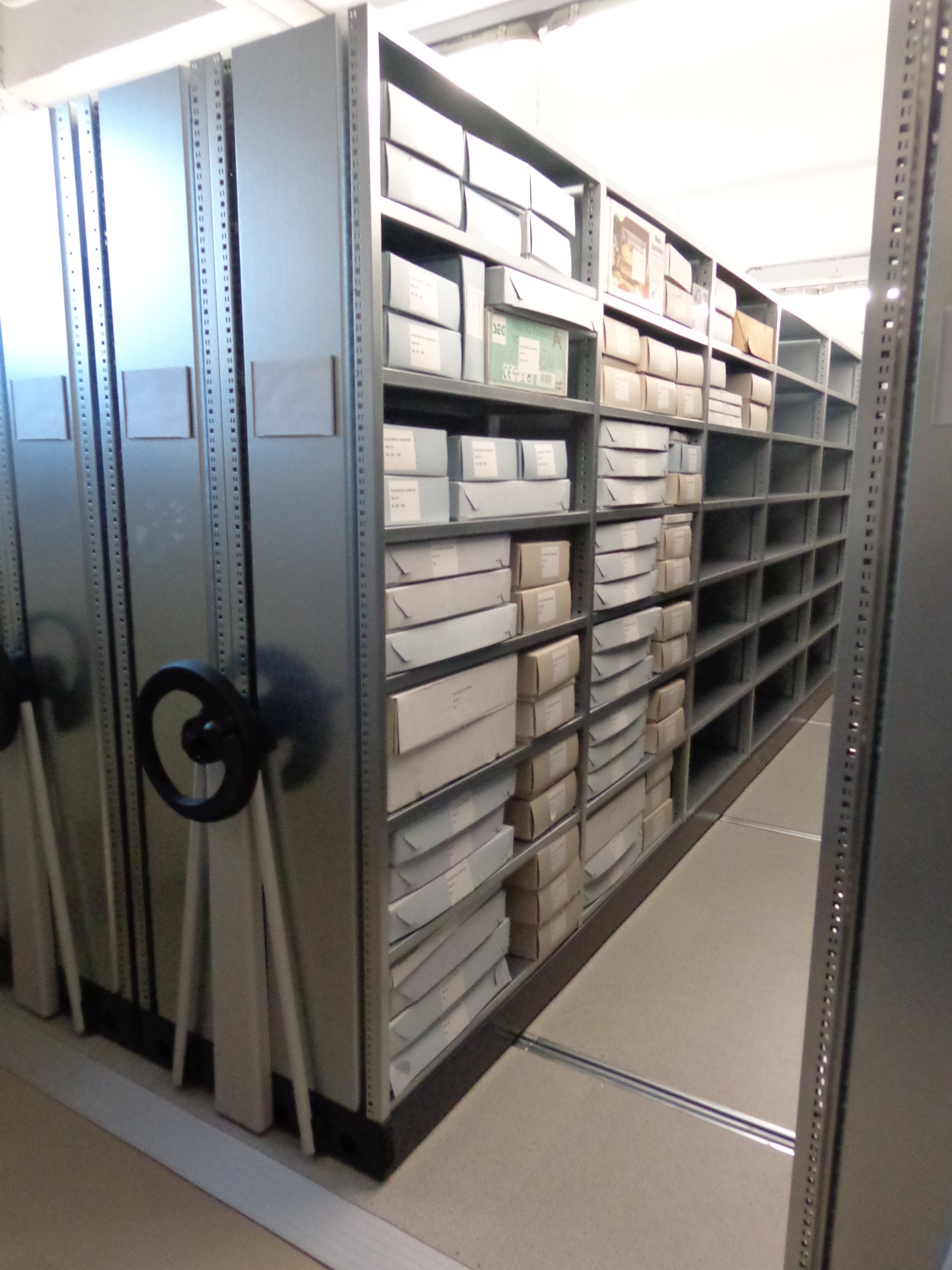 Blick durch einen Gang zwischen verschiebbaren Archivregalen. Die Metallregale sind überwiegend mit weißen Archivkartons unterschiedlicher Größe gefüllt, vereinzelt stehen weitere Schachteln und Materialien in den Fächern. Links sind die Handräder der Rollregalanlage zu sehen, der Gang führt in den hinteren Bereich des Magazins.