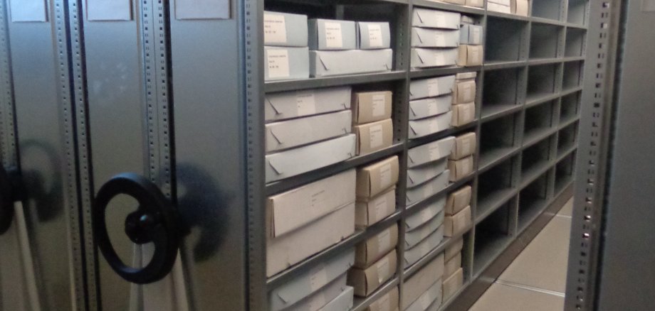 Stadtarchiv Lahnstein Blick durch einen Gang zwischen verschiebbaren Archivregalen. Die Metallregale sind überwiegend mit weißen Archivkartons unterschiedlicher Größe gefüllt, vereinzelt stehen weitere Schachteln und Materialien in den Fächern. Links sind die Handräder der Rollregalanlage zu sehen, der Gang führt in den hinteren Bereich des Magazins.