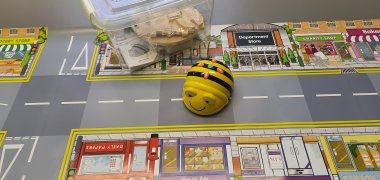 Bee-Bot-Lernroboter auf einer Spielmatte mit Straßen und Gebäuden, daneben eine Box mit Zubehör.