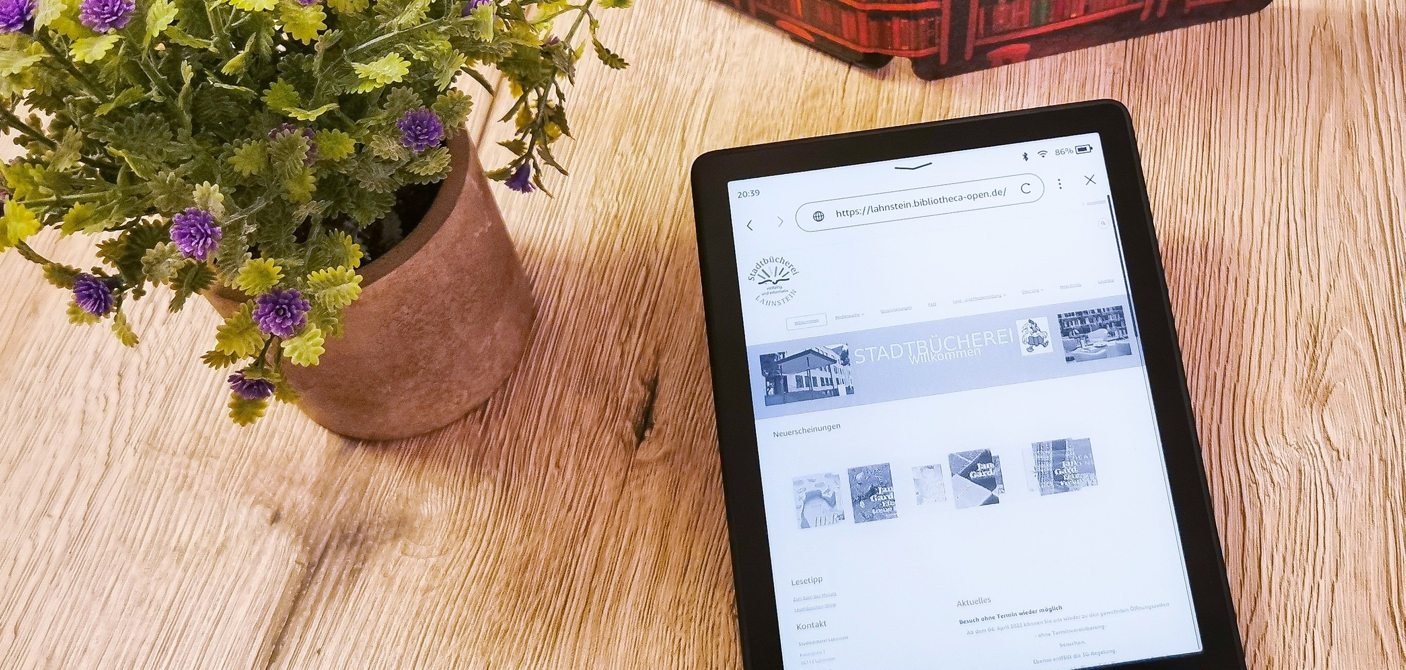 Auf einem Tisch befinden sich ein geöffneter eBook-Reader, ein aufgestelltes Buch und eine Blume