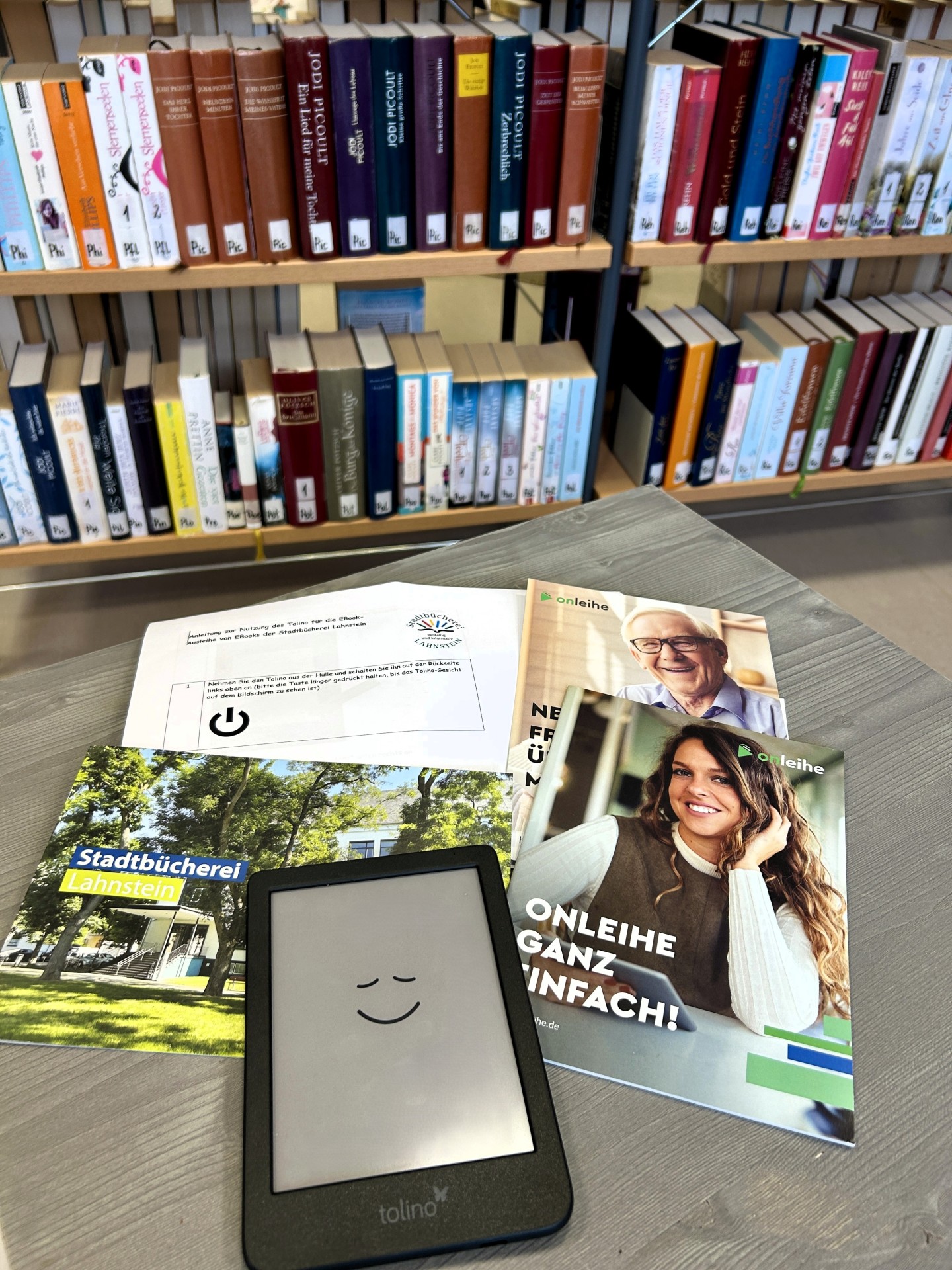 Auf einem Tisch in der Stadtbücherei Lahnstein liegen ein Tolino-eReader, Informationsbroschüren zur Onleihe und eine Kurzanleitung. Im Hintergrund sind mehrere Bücherregale mit Romanen und Sachbüchern zu sehen.