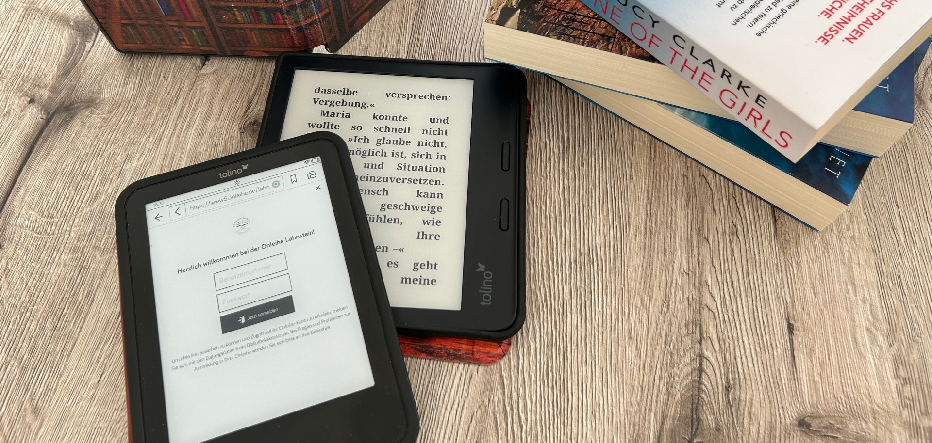 Auf einem Tisch befinden sich zwei ebook-Reader, im Hintergrund ein aufstellter eBook-Reader mit einer Hülle, die viele Bücherreihen zeigt, sowie ein Stapel von mehreren Büchern