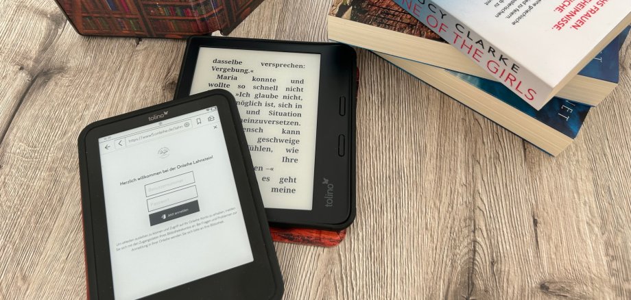 Auf einem Tisch befinden sich zwei ebook-Reader, im Hintergrund ein aufstellter eBook-Reader mit einer Hülle, die viele Bücherreihen zeigt, sowie ein Stapel von mehreren Büchern