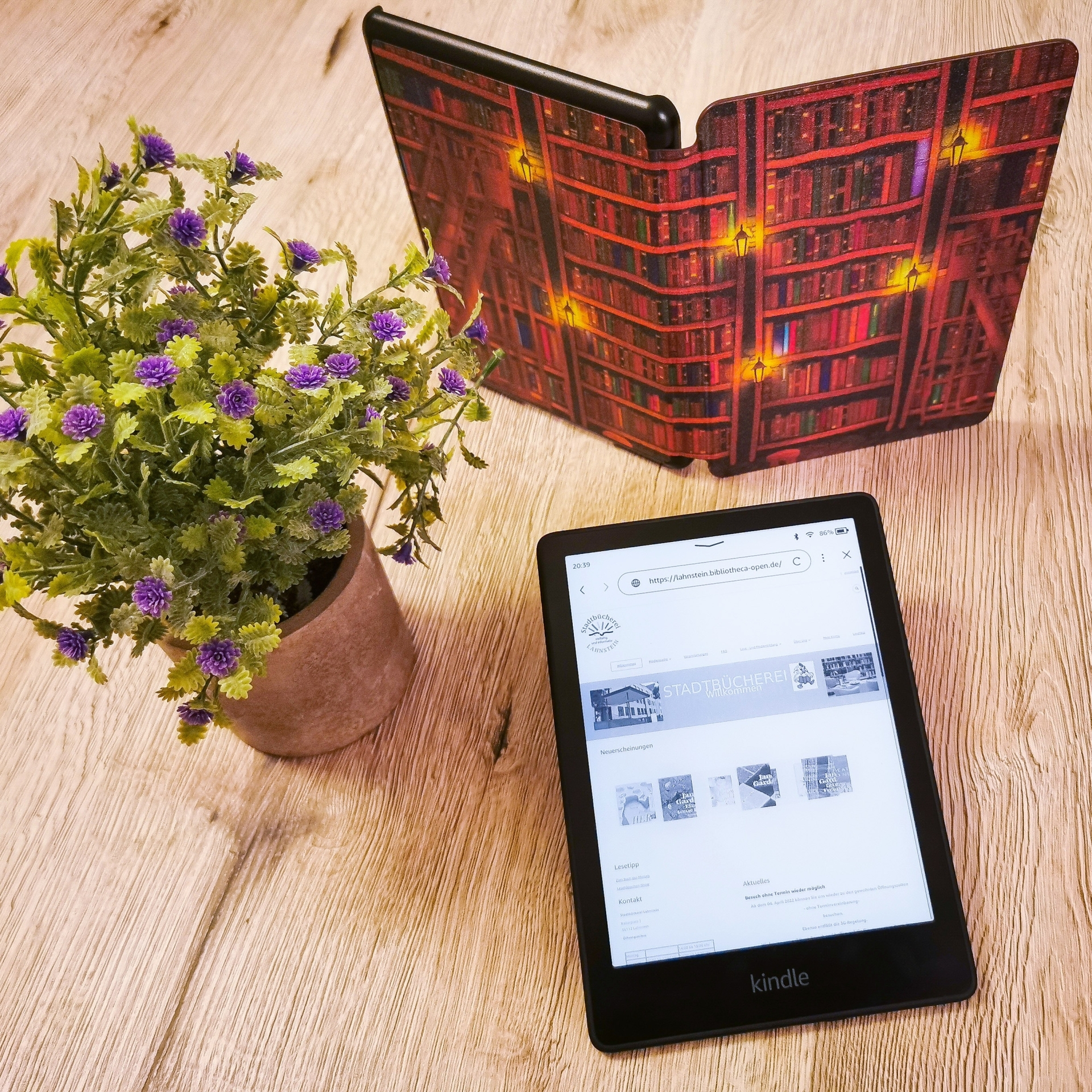 Auf einem Tisch befinden sich ein geöffneter eBook-Reader, ein aufgestelltes Buch und eine Blume