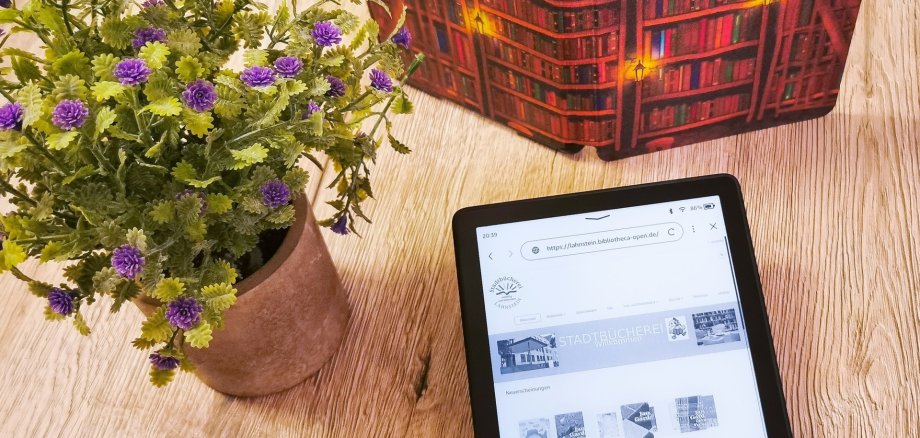 Stadtbücherei Lahnstein Auf einem Tisch befinden sich ein geöffneter eBook-Reader, ein aufgestelltes Buch und eine Blume