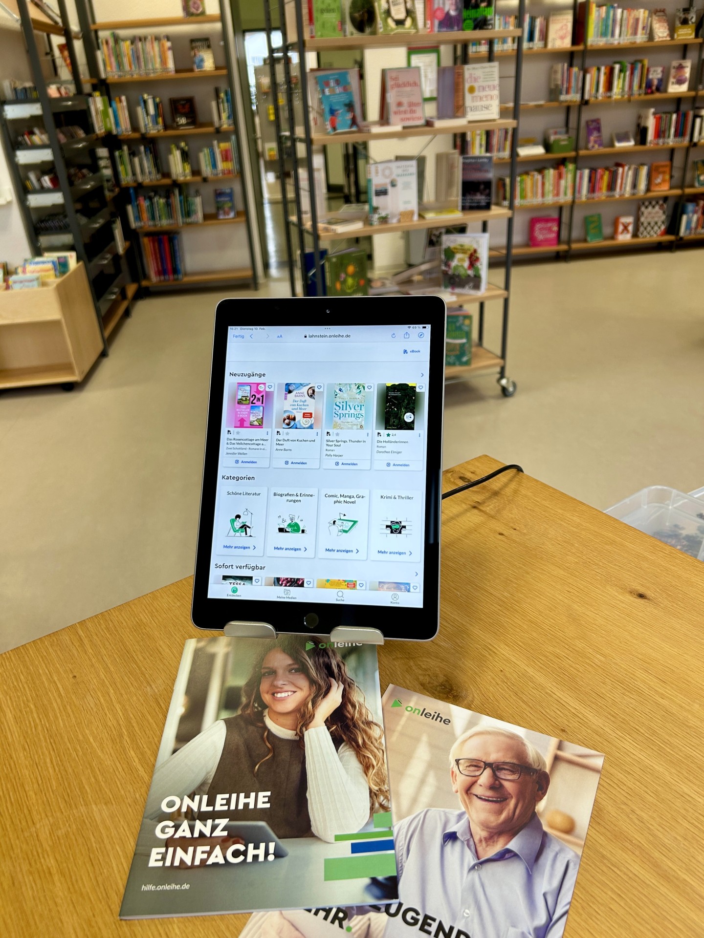 Ein Tablet zeigt die Startseite der Onleihe mit verschiedenen eBook-Angeboten. Vor dem Gerät liegen Informationsbroschüren zur digitalen Ausleihe. Im Hintergrund sind die Regale und Buchbestände der Stadtbücherei Lahnstein erkennbar.