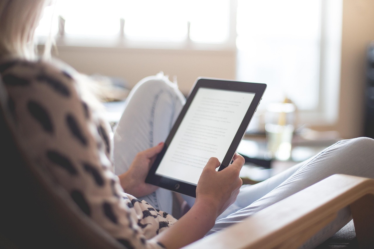 Eine Frau sitzt auf einer Couch und ist schräg von hinten abgelichtet. Sie hält einen eBook-Reader in der Hand.