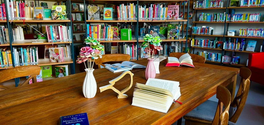 Stadtbücherei Lahnstein Ein heller Lesebereich in der Stadtbücherei mit einem großen Holztisch in der Bildmitte. Auf dem Tisch liegen mehrere aufgeschlagene Bücher sowie ein geschlossenes Buch, daneben stehen kleine Vasen mit dekorativen Papierblumen und eine hölzerne Vogelfigur. Rund um den Tisch befinden sich Holzstühle mit grauen Sitzpolstern. Im Hintergrund erstrecken sich hohe, gut gefüllte Bücherregale mit Romanen, Sachbüchern und Kochbüchern; einige Bücher werden frontal präsentiert. Rechts ist ein roter Sessel zu sehen, auf einem Regal steht ein Globus. Die freundliche und ruhige Atmosphäre lädt zum Lesen und Verweilen ein.