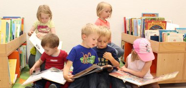 Mehrere Kinder sitzen in der Stadtbücherei Lahnstein und lesen zusammen. Auf beiden Seiten stehen niedrige Holzkosten, die mit Büchern gefüllt sind.