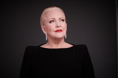 Angelika Milster Porträt einer elegant gekleideten Person mit kurzem, zurückgekämmtem, hellblondem Haar. Sie trägt ein schwarzes Oberteil und lange, funkelnde Ohrringe. Ihr Make-up ist dezent mit betonten roten Lippen. Die Person blickt leicht nach oben zur Seite, vor einem dunklen, neutralen Hintergrund.