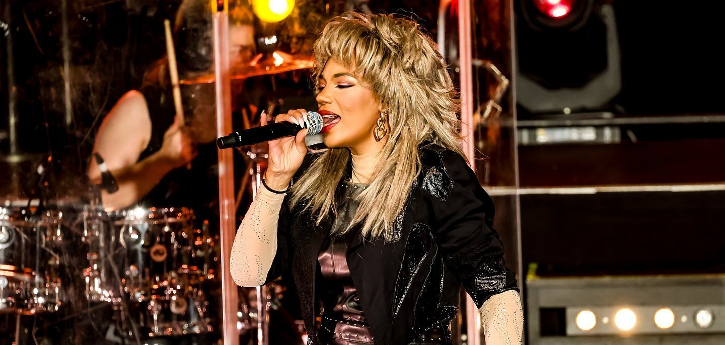 Eine Sängerin mit voluminöser blonder Frisur steht auf einer Bühne und singt in ein Mikrofon. Sie trägt ein schwarzes, glänzendes Outfit mit langen, hellen Ärmeln und auffälligen Ohrringen. Im Hintergrund ist ein Schlagzeuger hinter einer transparenten Schutzwand zu sehen, beleuchtet von warmem Bühnenlicht.