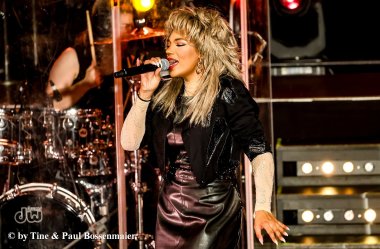 TINA – The Rock Legend“ Eine Sängerin mit voluminöser blonder Frisur steht auf einer Bühne und singt in ein Mikrofon. Sie trägt ein schwarzes, glänzendes Outfit mit langen, hellen Ärmeln und auffälligen Ohrringen. Im Hintergrund ist ein Schlagzeuger hinter einer transparenten Schutzwand zu sehen, beleuchtet von warmem Bühnenlicht.