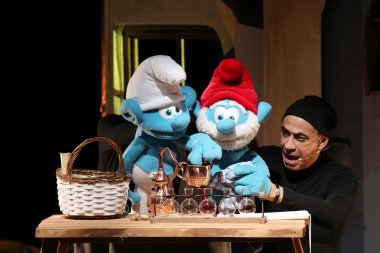 Die Schlümpfe - das Musical Zwei große Schlumpf-Figuren stehen auf einer Theaterbühne an einem Tisch mit kleinen Glasgefäßen und Kupferkannen. Einer der Schlümpfe trägt eine rote Mütze und einen weißen Bart, der andere eine große weiße Mütze. Rechts daneben spielt ein Darsteller in schwarzer Kleidung die Szene mit und spricht mit geöffnetem Mund. Auf dem Tisch stehen außerdem ein geflochtener Korb und verschiedene Requisiten.