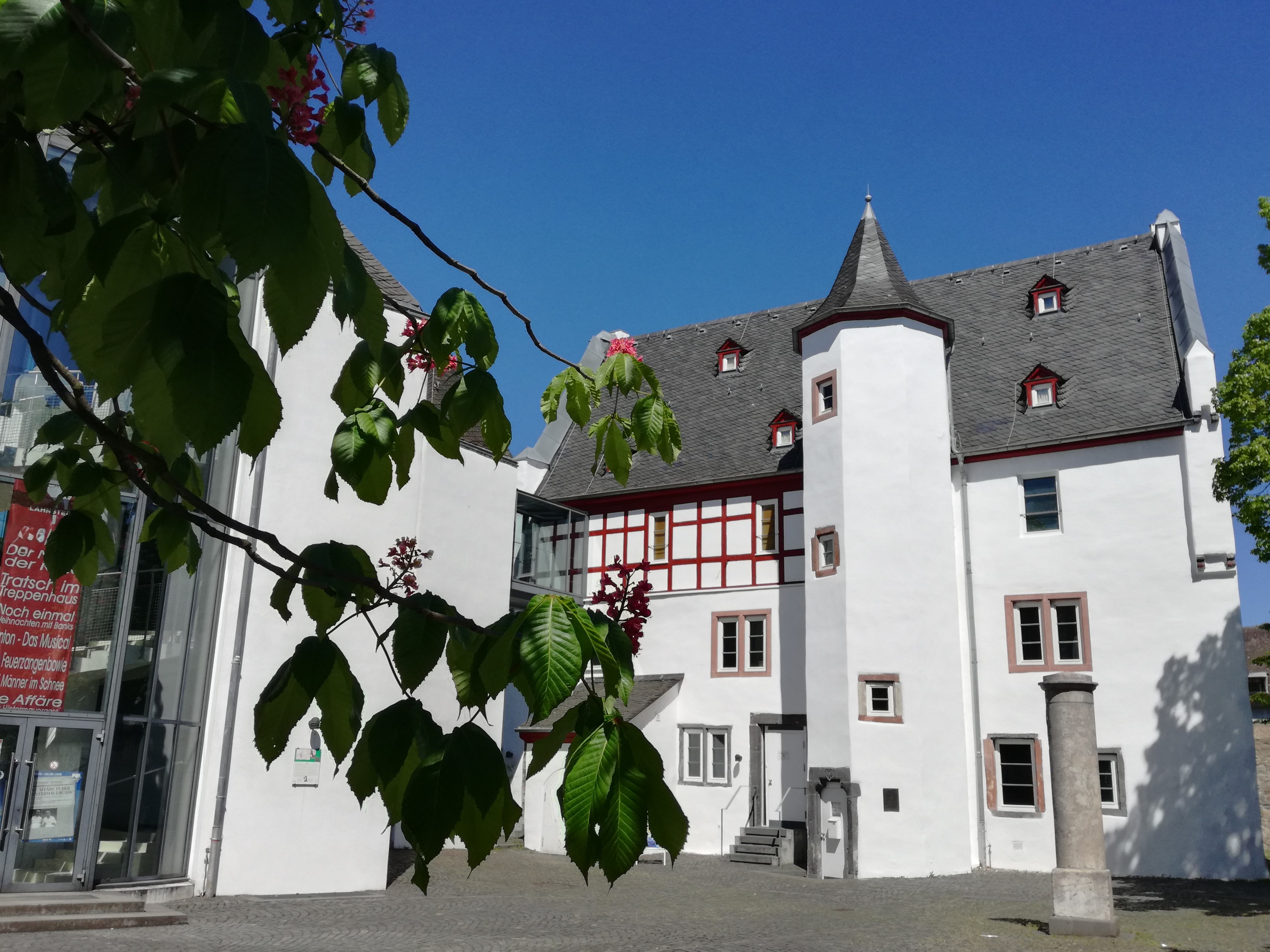 Außenansicht eines historischen Gebäudekomplexes mit weiß verputzten Fassaden und dunklen Schieferdächern unter strahlend blauem Himmel mit grünen Blättern im Vordergrund.