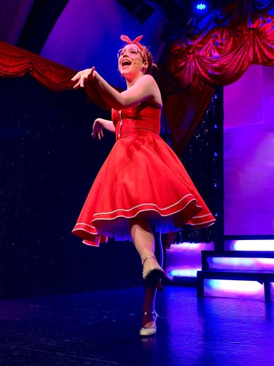 Eine Darstellerin in einem roten Kleid und mit rotem Haarschmuck steht auf einer beleuchteten Theaterbühne. Sie hebt ein Bein an und streckt einen Arm nach vorne. Der Bühnenbereich ist in violettes und blaues Licht getaucht.
