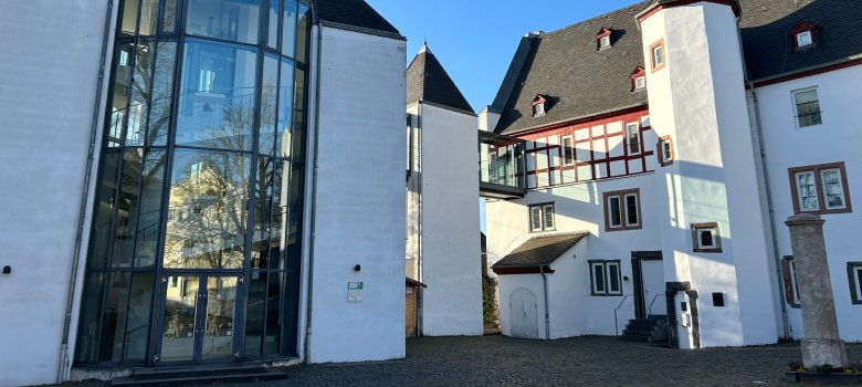 Außenansicht eines historischen Gebäudekomplexes mit weiß verputzten Fassaden und dunklen Schieferdächern unter strahlend blauem Himmel. Links befindet sich ein moderner gläserner Anbau mit hoher Fensterfront, rechts ein Gebäudeteil mit Fachwerkelementen und einem kleinen Turm. Der gepflasterte Innenhof liegt im Sonnenlicht.