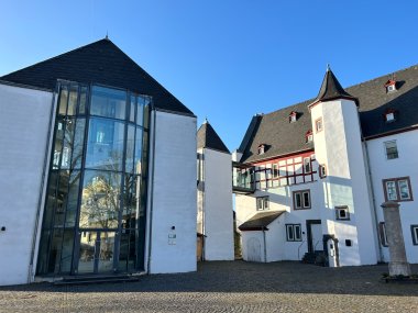Außenansicht eines historischen Gebäudekomplexes mit weiß verputzten Fassaden und dunklen Schieferdächern unter strahlend blauem Himmel. Links befindet sich ein moderner gläserner Anbau mit hoher Fensterfront, rechts ein Gebäudeteil mit Fachwerkelementen und einem kleinen Turm. Der gepflasterte Innenhof liegt im Sonnenlicht.