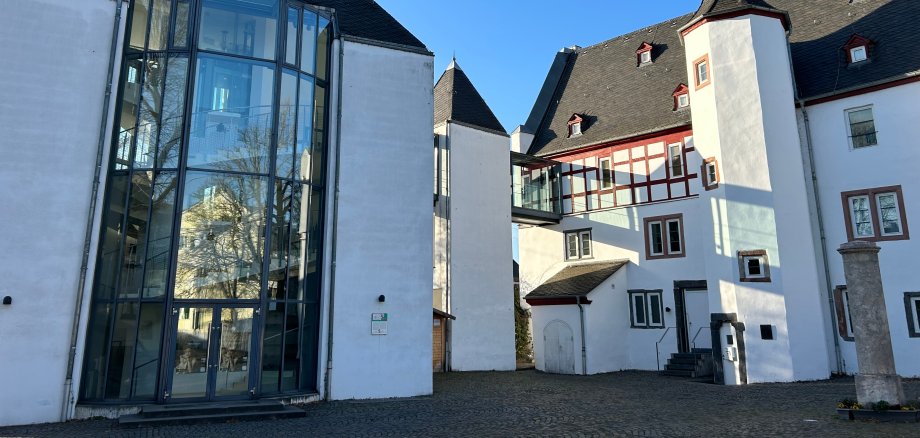 Theater Lahnstein Außenansicht eines historischen Gebäudekomplexes mit weiß verputzten Fassaden und dunklen Schieferdächern unter strahlend blauem Himmel. Links befindet sich ein moderner gläserner Anbau mit hoher Fensterfront, rechts ein Gebäudeteil mit Fachwerkelementen und einem kleinen Turm. Der gepflasterte Innenhof liegt im Sonnenlicht.