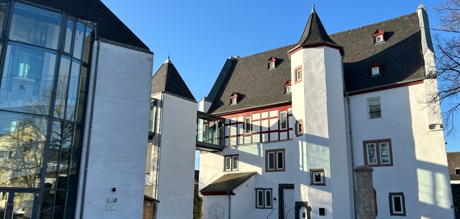 Theater Lahnstein Außenansicht eines historischen Gebäudekomplexes mit weiß verputzten Fassaden und dunklen Schieferdächern unter strahlend blauem Himmel. Links befindet sich ein moderner gläserner Anbau mit hoher Fensterfront, rechts ein Gebäudeteil mit Fachwerkelementen und einem kleinen Turm. Der gepflasterte Innenhof liegt im Sonnenlicht.