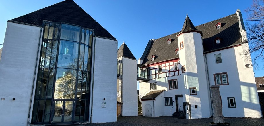 Theater Lahnstein Außenansicht eines historischen Gebäudekomplexes mit weiß verputzten Fassaden und dunklen Schieferdächern unter strahlend blauem Himmel. Links befindet sich ein moderner gläserner Anbau mit hoher Fensterfront, rechts ein Gebäudeteil mit Fachwerkelementen und einem kleinen Turm. Der gepflasterte Innenhof liegt im Sonnenlicht.