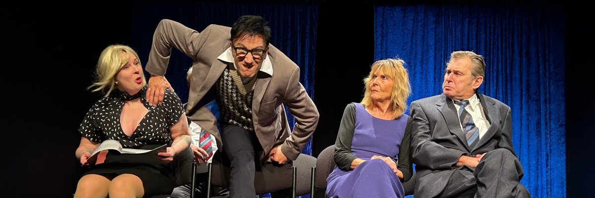 Vier Personen sitzen nebeneinander auf Stühlen auf einer Theaterbühne. Ein Mann im Anzug beugt sich zwischen zwei der Sitzenden nach vorne. Die Szene wird vor einem blauen Vorhang gespielt.