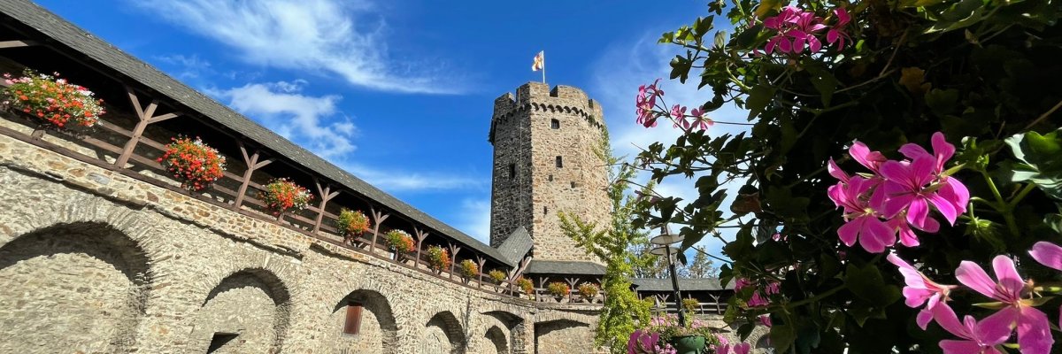 Hexenturm Lahnstein mit Stadtmauer Historischer steinerner Turm, der Hexenturm. Im Vordergrund blüht ein Strauch mit rosa Blüten, dahinter ist ein blauer Himmel zu sehen.