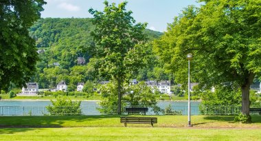 Rheinanlagen Oberlahnstein Grüne Parkanlage mit Rasenfläche, mehreren Bäumen und einer Bank. Im Hintergrund verläuft ein Weg entlang des Flusses Rhein.