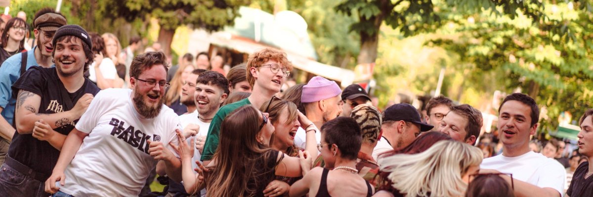 Lahneck Live Festival in Lahnstein Eine Gruppe junger Festivalbesucher tanzt und bewegt sich ausgelassen vor der Bühne. Die Menschen lachen, springen und feiern gemeinsam unter großen Bäumen im Grünen. Die sommerliche Stimmung vermittelt die Energie eines lebendigen Open-Air-Konzerts.