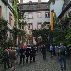Führung zum CCO-Fastnachtsmuseum in Schloss Martinsburg Lahnstein Mehrere Menschen stehen in einem begrünten Innenhof vor einem historischen Gebäude. Die Gruppe scheint einer Führung oder Präsentation zu folgen.