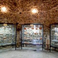 Ehemaliges Verlies im Hexenturm Lahnstein mit heutigem Heimatmuseum Innenraum eines historischen Gebäudes mit Steinmauern und Holzbalkendecke. Der Raum ist durch mehrere Wandlampen beleuchtet. An der Wand stehen Vitrinen mit historischen Ausstellungsstücken.
