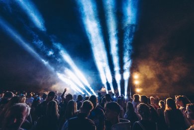 Rhein in Flammen Lahnstein Menschen stehen dicht zusammen vor einer Open-Air-Bühne und feiern ein Konzert bei Nacht. Helle Scheinwerferstrahlen und Nebel durchbrechen den dunklen Himmel, während ein DJ oder Künstler auf der Bühne auftritt und die Lichtshow die Festivalatmosphäre prägt.